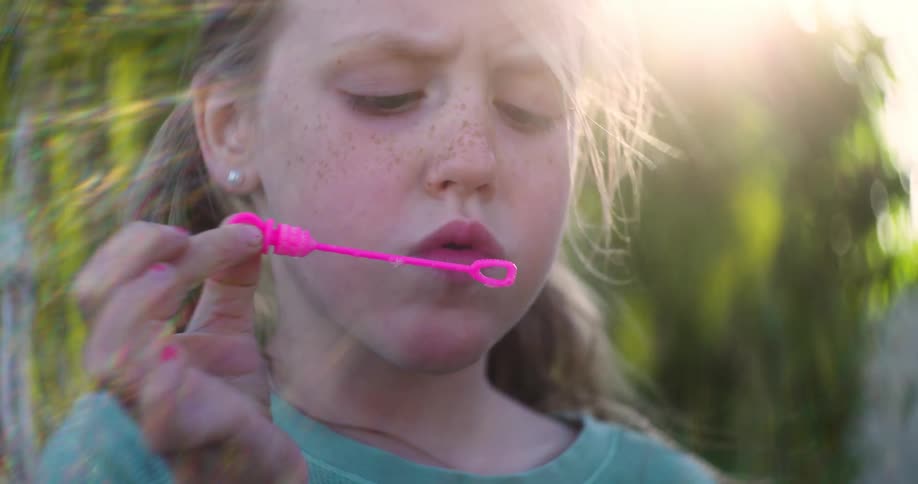 Close up of a young girl blowing bubbles from a pink wand outdoors