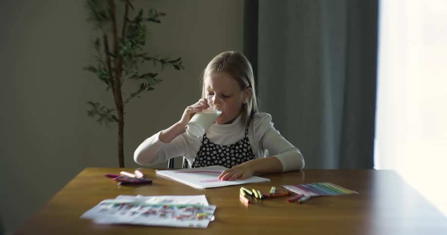 A young girl takes a break from drawing to drink from a glass of milk