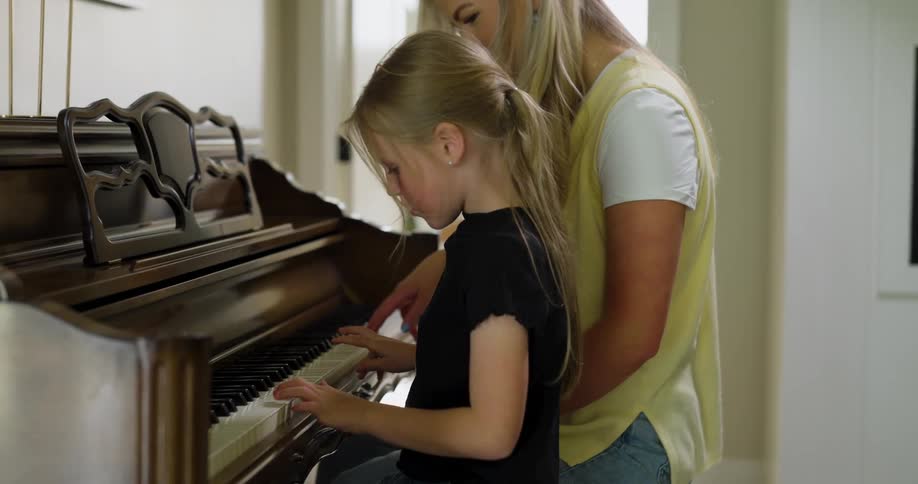An adult woman next to a little blonde girl is helping her play a piano
