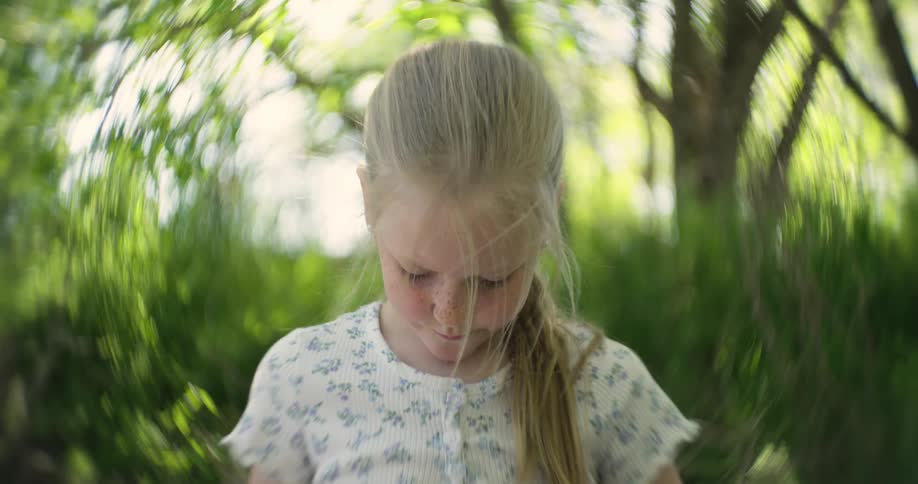 Little blonde girl looking down while standing in natural surroundings with trees around