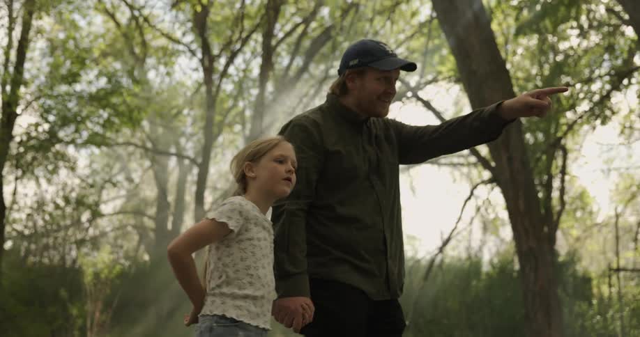 Father and daughter walk on a park path surrounded by trees and the father points ahead
