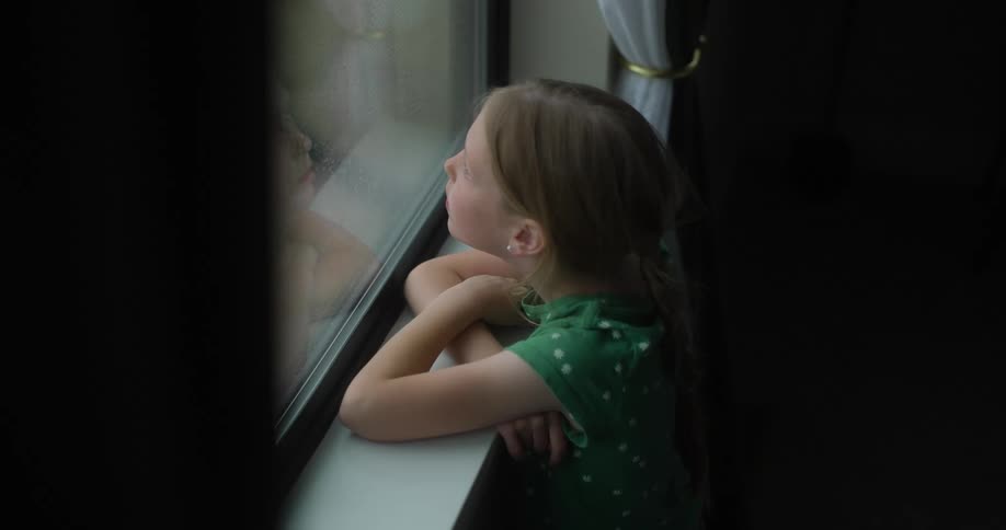 A little girl in a green dress is looking out the window at the rain