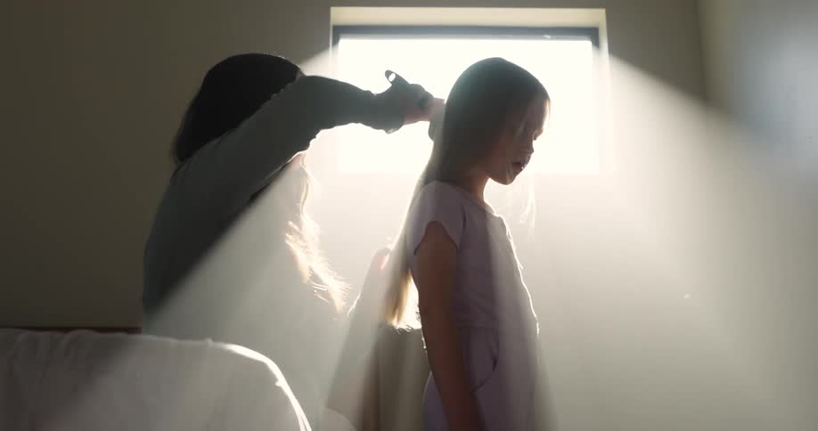 A mother brushes the long hair of her daughter as sunlight shines through the window
