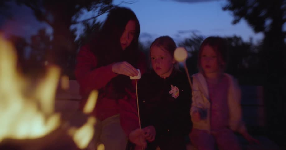 Mother and two little girls put marshmallows on sticks to hold over a camp fire