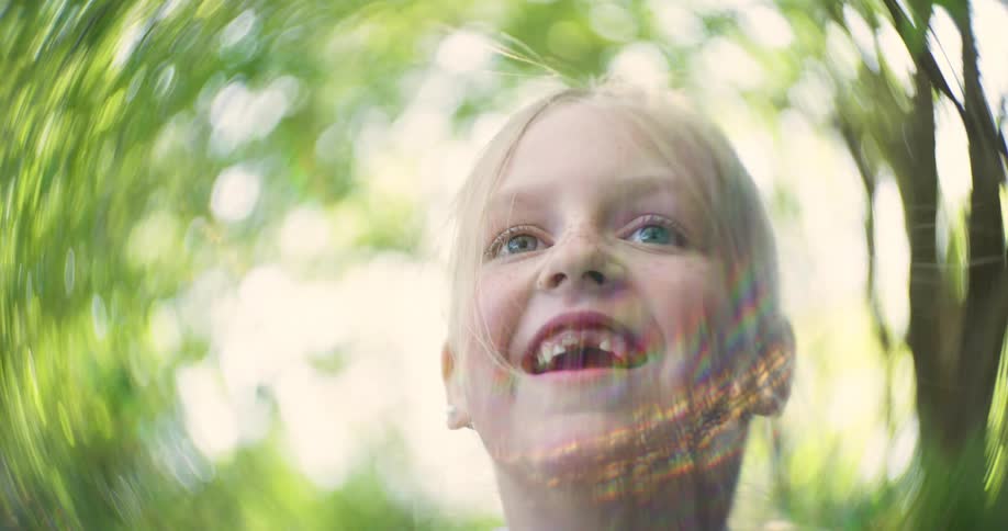 The sun casts a colorful rainbow effect around the face of a young girl yelling in a forest