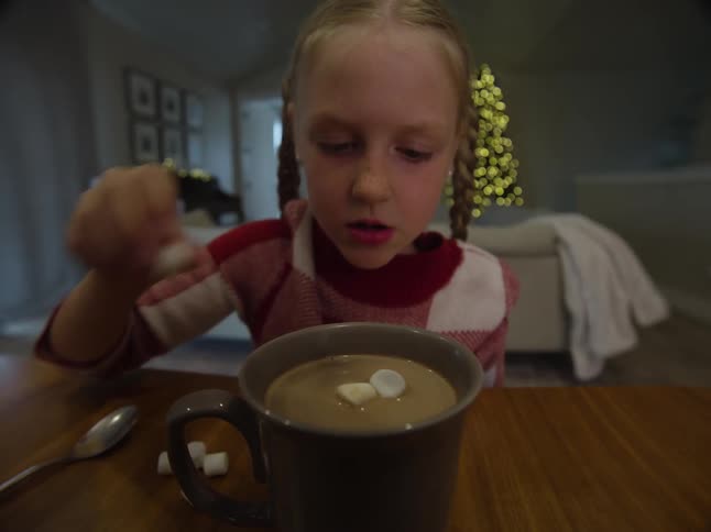 Young blonde girl in pig tails puts marshmallows into a cup of hot chocolate