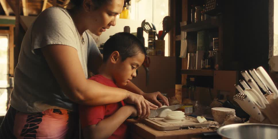 Mother guiding her sons hands to cut vegetables with a knife on a wooden cutting board