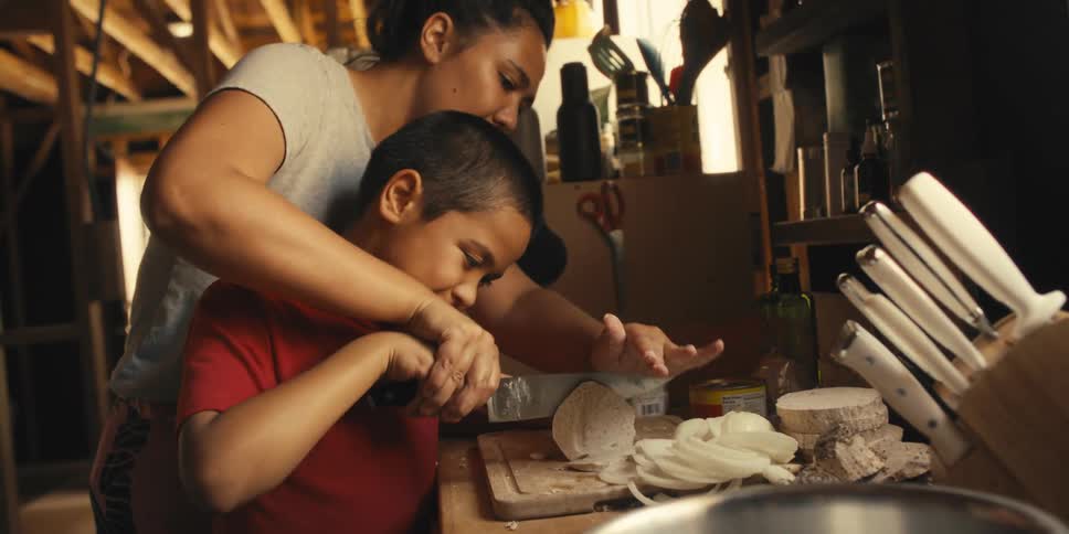 Mother and son together handling a knife making a cut in food on a wooden board