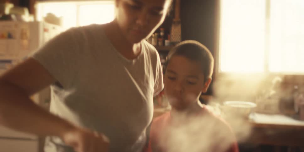 Son stands in the kitchen and watches while his mother stirs food in a pot on the stove