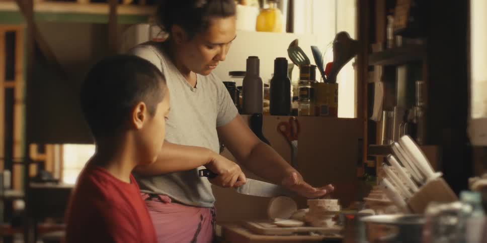 A woman is using a knife to cut something next to a boy who is watching
