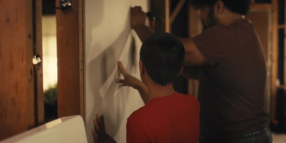 Boy holding a sheet of drywall while his father next to him uses a nail and a hammer