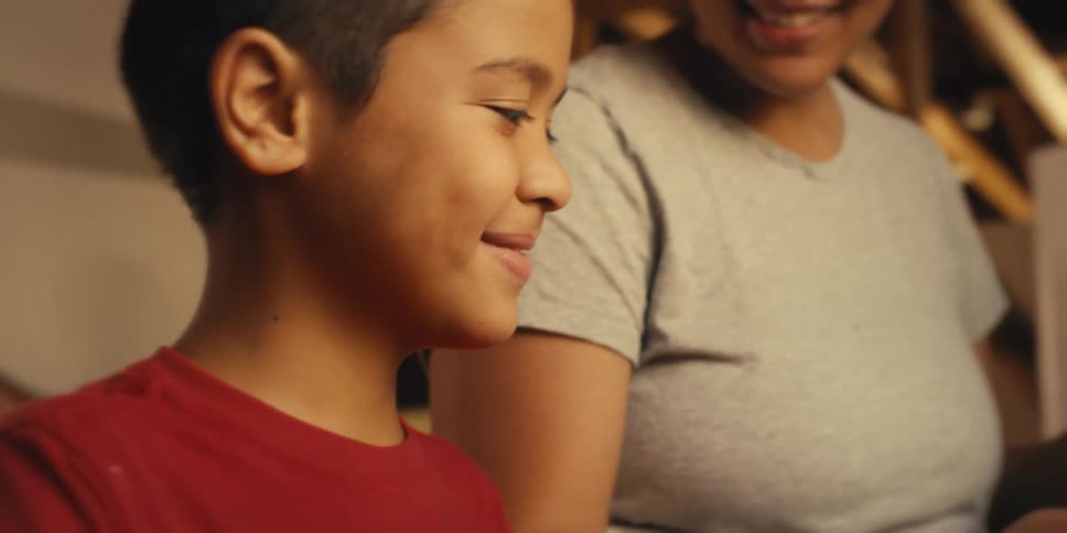 A young boy smiles while working on a project with an adult in the background