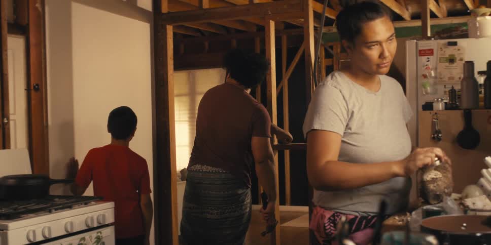 A mother prepares a meal as her husband and son put up drywall