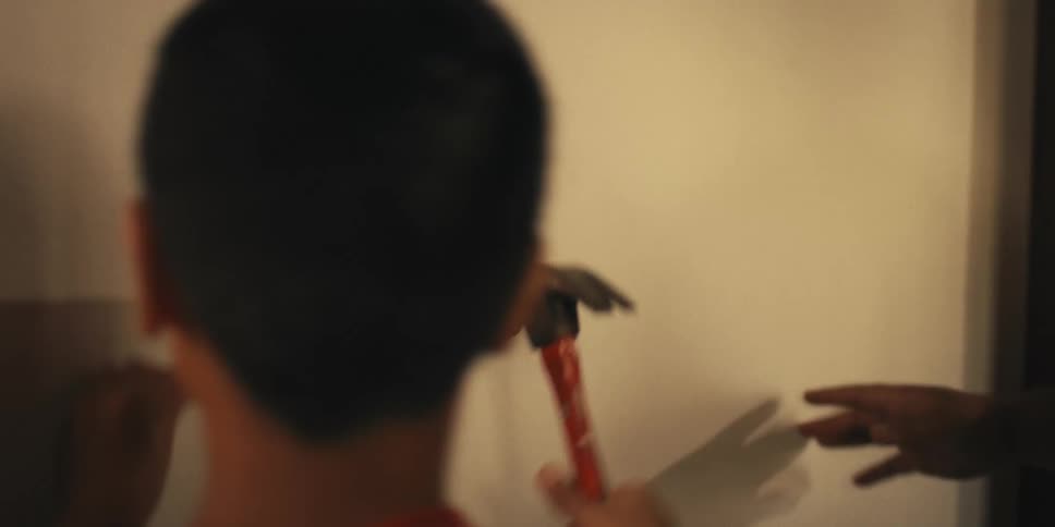 A man hands a nail and a hammer to a boy standing next to him and the boy hammers the nail into a wall