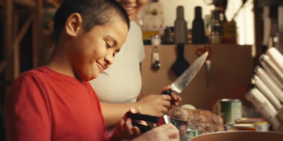 Mother holding a kitchen knife smiles while watching her son open a can