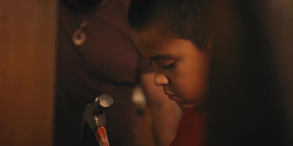 Young boy is handed a hammer and nail and begins to drive it into the wall