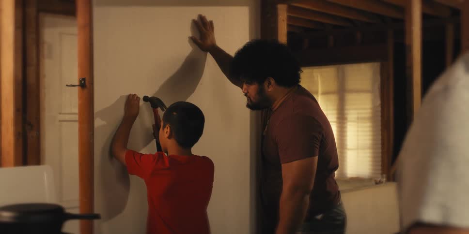 Father guiding his young son on how to use a hammer on a piece of sheet rock as a woman peels a root in the foreground