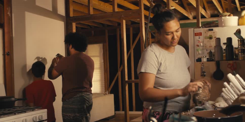 A mother prepares food in a partially built kitchen while her son goes to watch his father drive a nail into a wall
