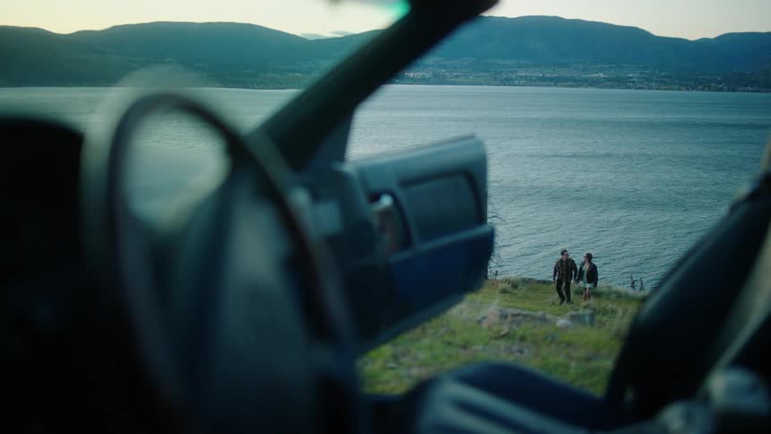 Couple holds hands while walking up a hill from the water toward a car with an open door