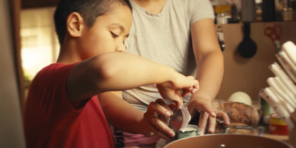 Boy next to his mother helping prepare a meal and pouring coconut milk into the pan