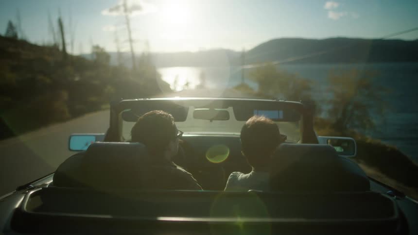 Two people in a convertible car on a seaside road with a sun on the horizon