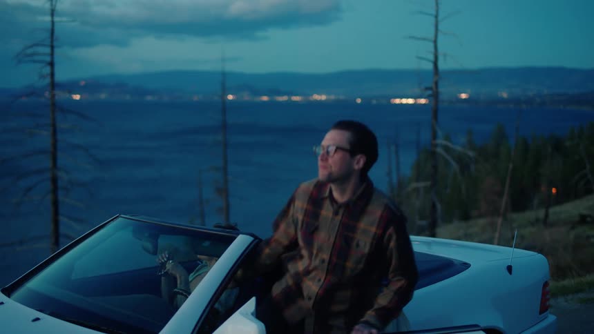 A man and woman share a romantic moment sitting on the side of a vehicle before getting in and driving off