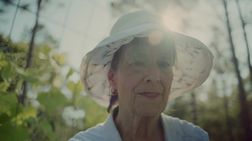 Elderly woman wearing a white sun hat standing in sunlight among greenery
