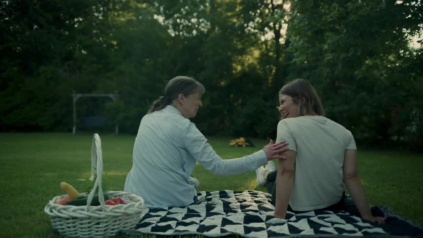 Two women sit on an outdoor blanket and watch a young girl play in the backyard