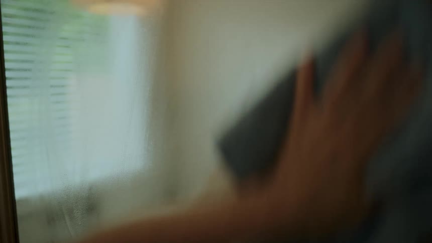 A woman using cleaning spray and cloth to clean a glass in bathroom