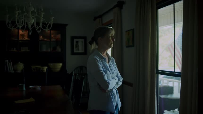 Teary eyed older woman rubs her arm while standing and staring out the window
