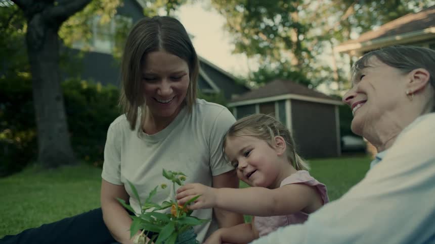 Three generations of females play with a flowering plant while sitting on the lawn