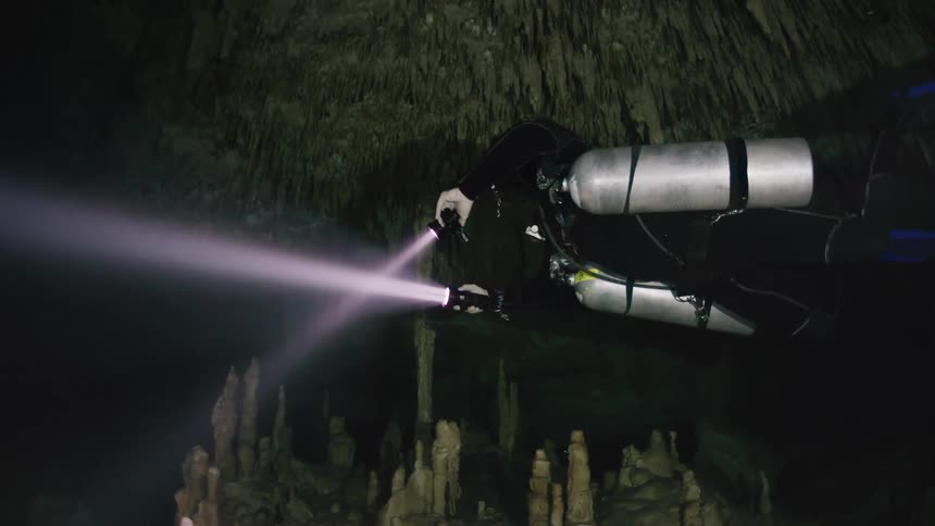 A diver underwater in a cave with stockpiles and he is holding two flashlights
