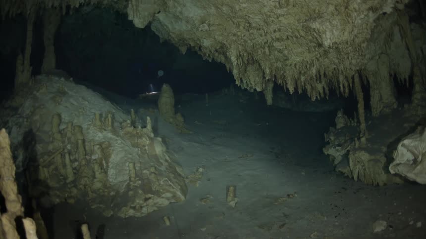 A scuba diver with a flashlight swims through a dark underwater cave