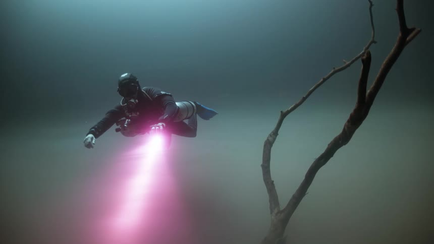 A scuba diver slowly swims underwater while shining a flashlight at the ocean floor
