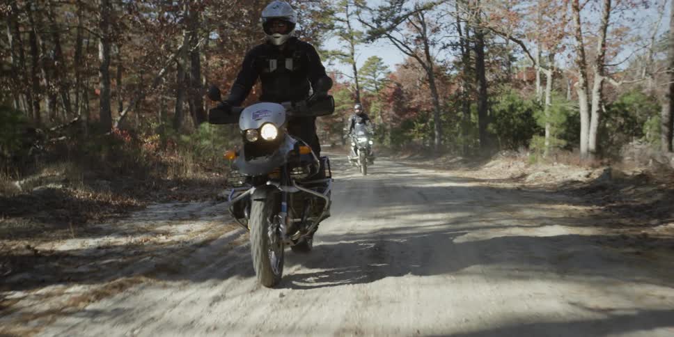 Two people ride motorcycles down a dirt road lined with trees