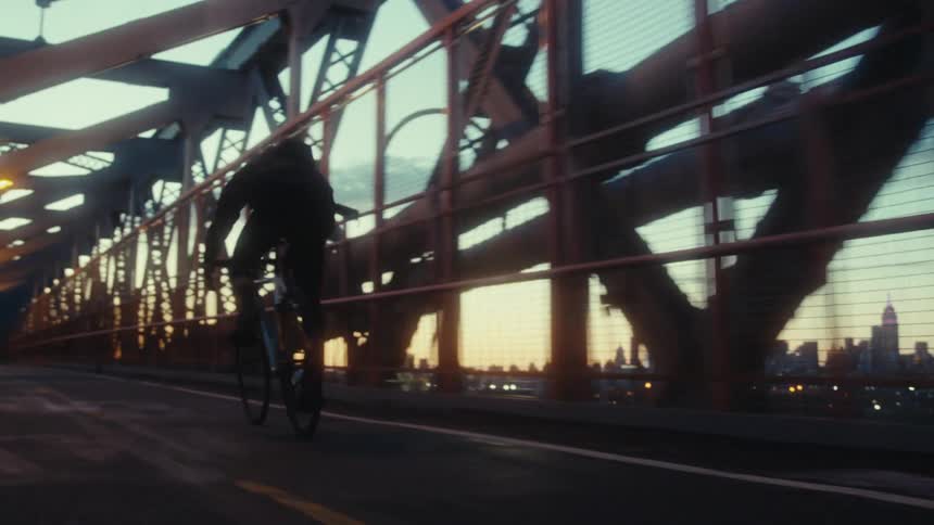 Cyclist rides across bridge over water with city skyline standing against a red horizon in the background