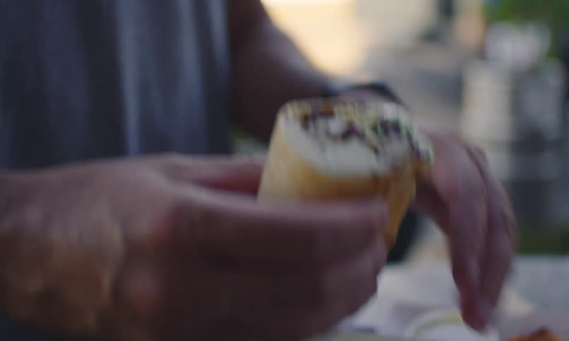 Man eating a sub sandwich at an outdoor table dips it in sauce
