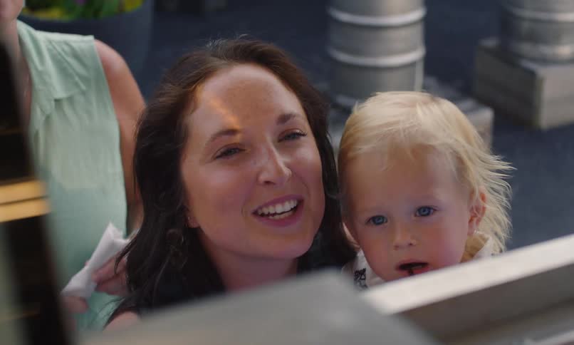 A woman at the front of a line outside a trailer ordering something while smiling and holding a baby