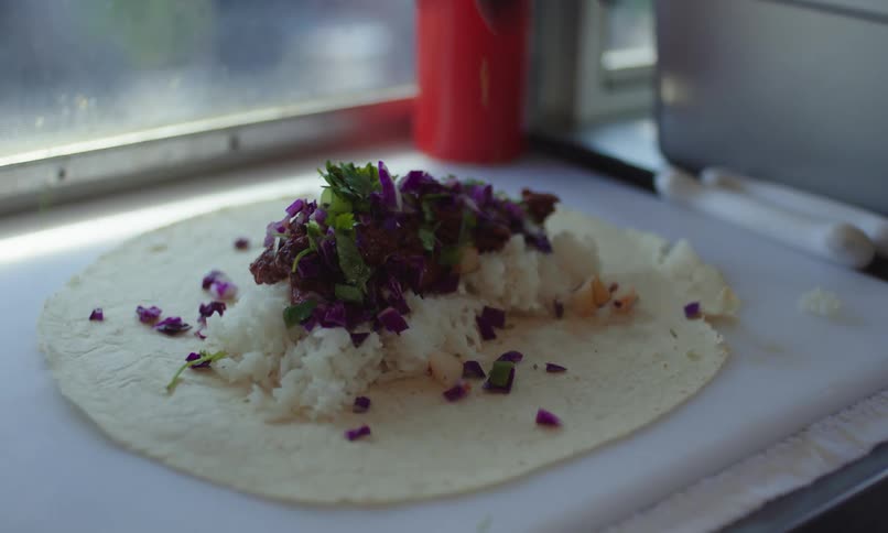 Gloved hand adding toppings to a burrito wrap with rice and vegetables
