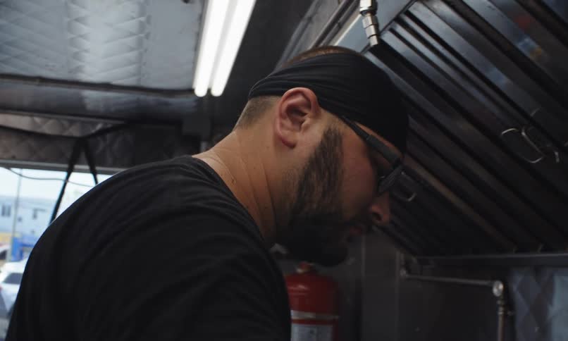 A man with glasses and wearing a black headband is preparing food and using a kitchen utensil to pick up the food