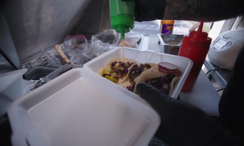 A man wearing a bandana and glasses is preparing food in a styrofoam container and putting a sauce on top