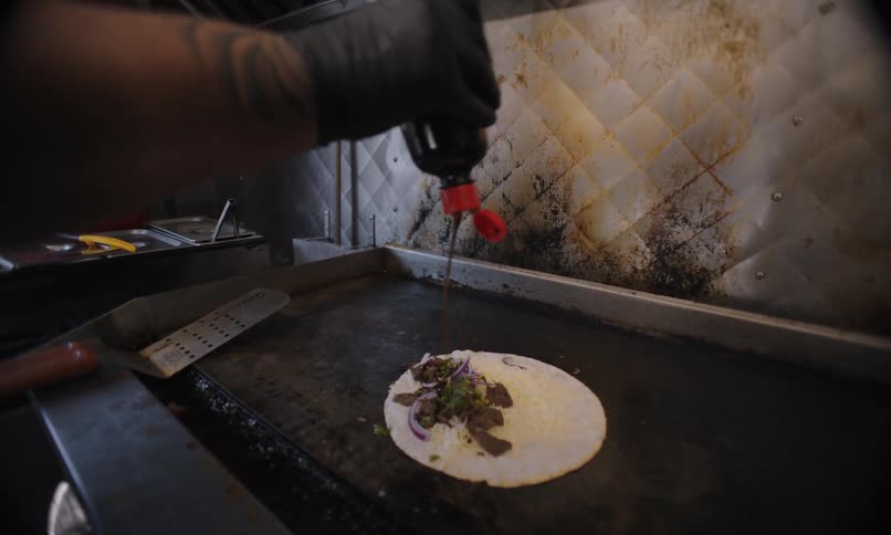Gloved hand adds red sauce to an open tortilla with ingredients on a metal cooking surface