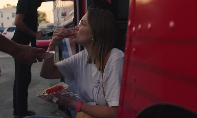 A woman sits on the step of a food truck and eats food