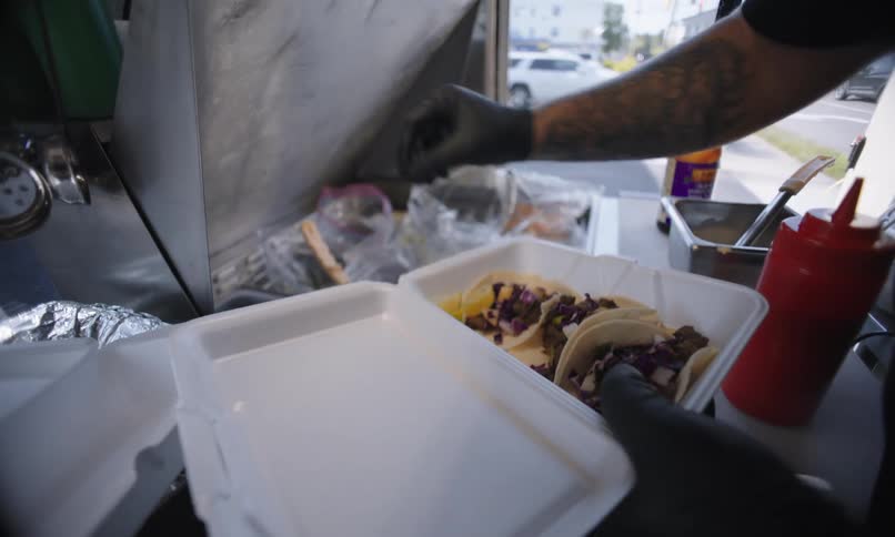 A man is wearing glasses and a cap on his head and he is preparing food on a styrofoam tray