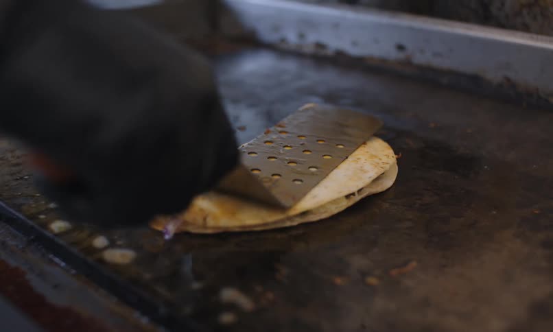 Hand with black gloves handles a metal spatula to cook a tortilla on a hot griddle