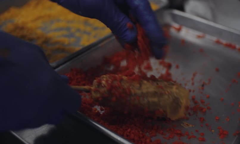 Gloved hands rolling a corndog in a tray of crushed red seasoning