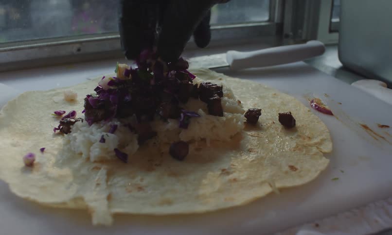 A persons gloved hand sprinkles beef onto a tortilla