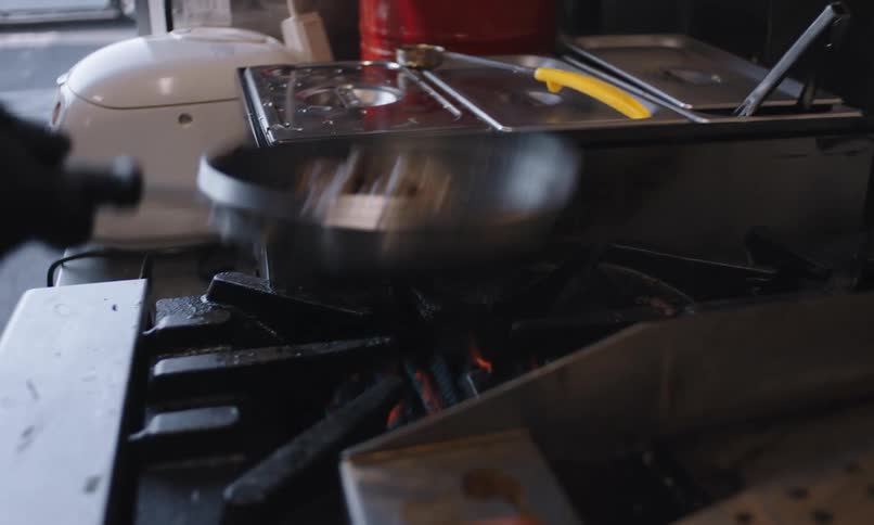 Human hands with black gloves holding a frying pan with meat that is frying on a stove