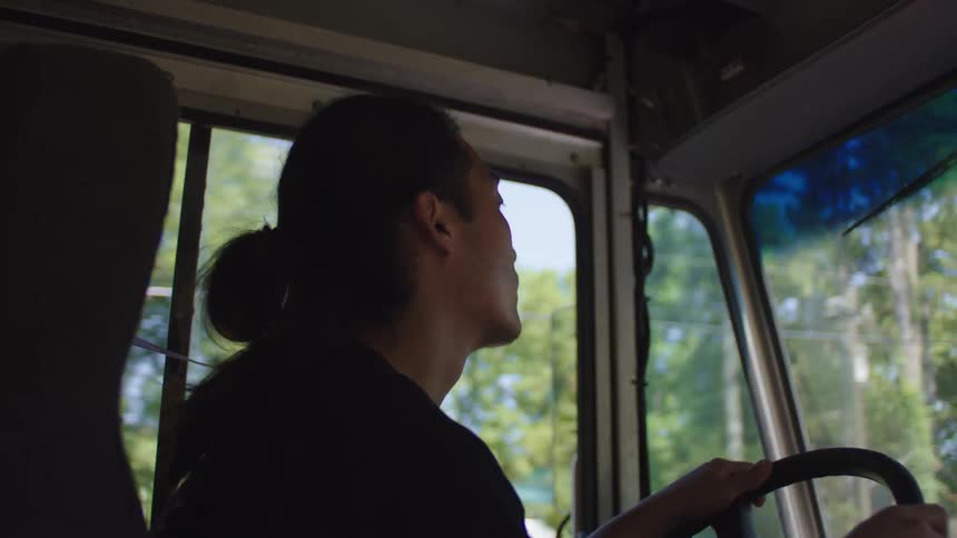 An asian man with a ponytail drives a large vehicle down a street
