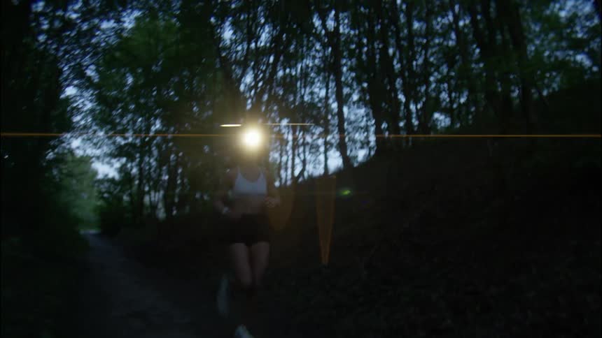 A woman running on a forest path at night with a lantern attached to her head